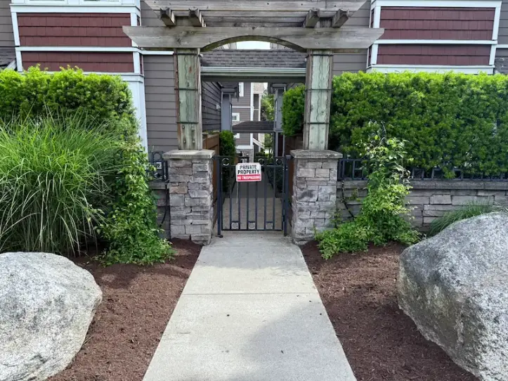 Walkway leading to a gated apartment entrance with stone pillars, wooden pergola arch, and landscaped greenery on both sides.